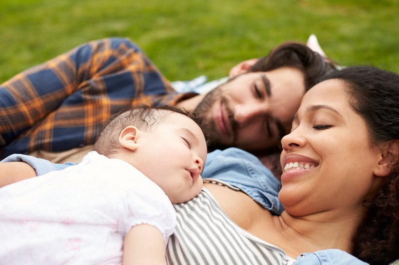 A multiracial couple, holding sleeping baby while outside on a picnic blanket